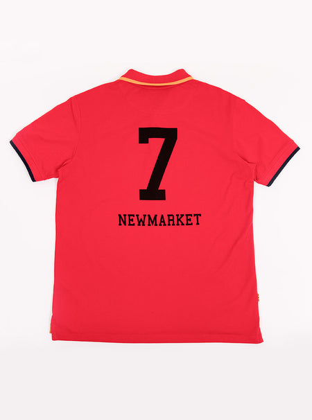 Flat lay of men's red polo shirt in 100% cotton with Newmarket 875 crest embroidered on the front with navy sleeve trimming and gold collar trimming.